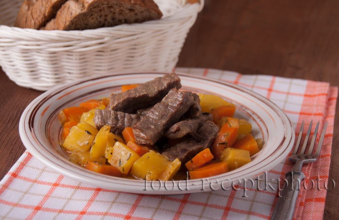 На фото тушеное мясо с овощами репы и моркови в тарелке, которая стоит на клетчатой салфетке и на заднем плане белая хлебница с черным хлебом