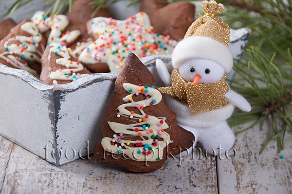 На фото сметанное печенье декорированное белым шоколадом и кондитерской посыпкой в деревянном ящичке и рядом игрушка снеговика и ветки ели