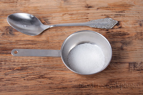 На фото сахарный песок в металлической кокотнице и столовая ложка на деревянном столе