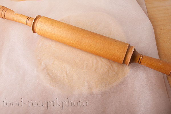 На фото песочное тесто на пекарской бумаге раскатываемое скалкой