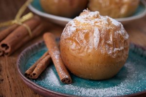На фото печеное яблоко с сахарной пудрой в бирюзовой тарелке и палочки корицы